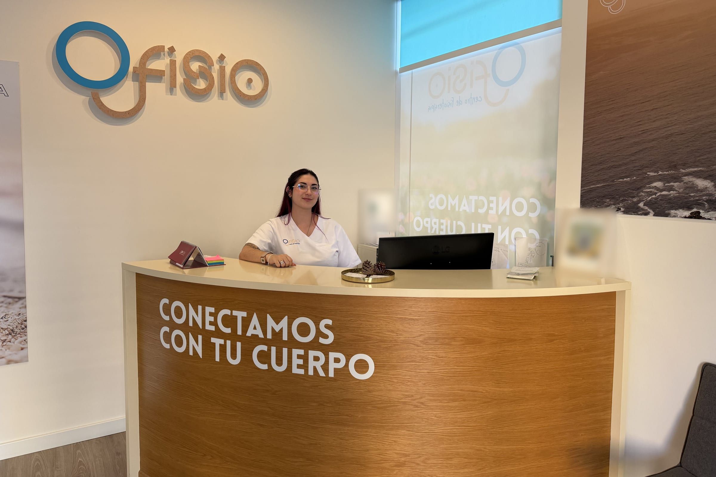Una profesional de la clínica sonríe tras el mostrador de recepción, con el lema «Conectamos con tu cuerpo» y el logo de Ofisio en la pared.